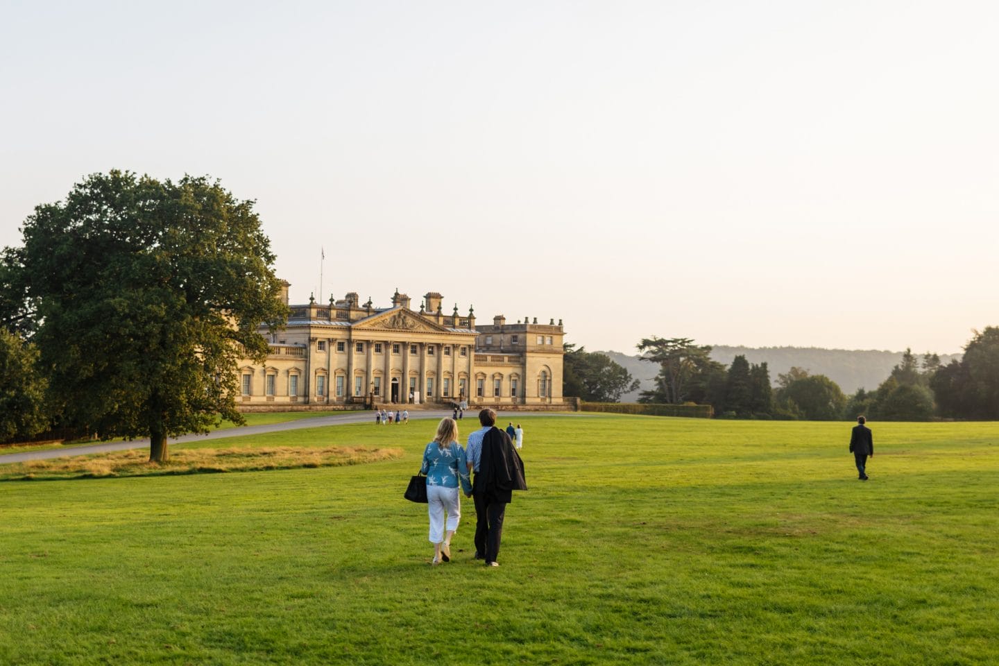 The golden glow of the setting sun bounces off the warm Palladian style country house, as people walk hand in hand across the large front lawn.