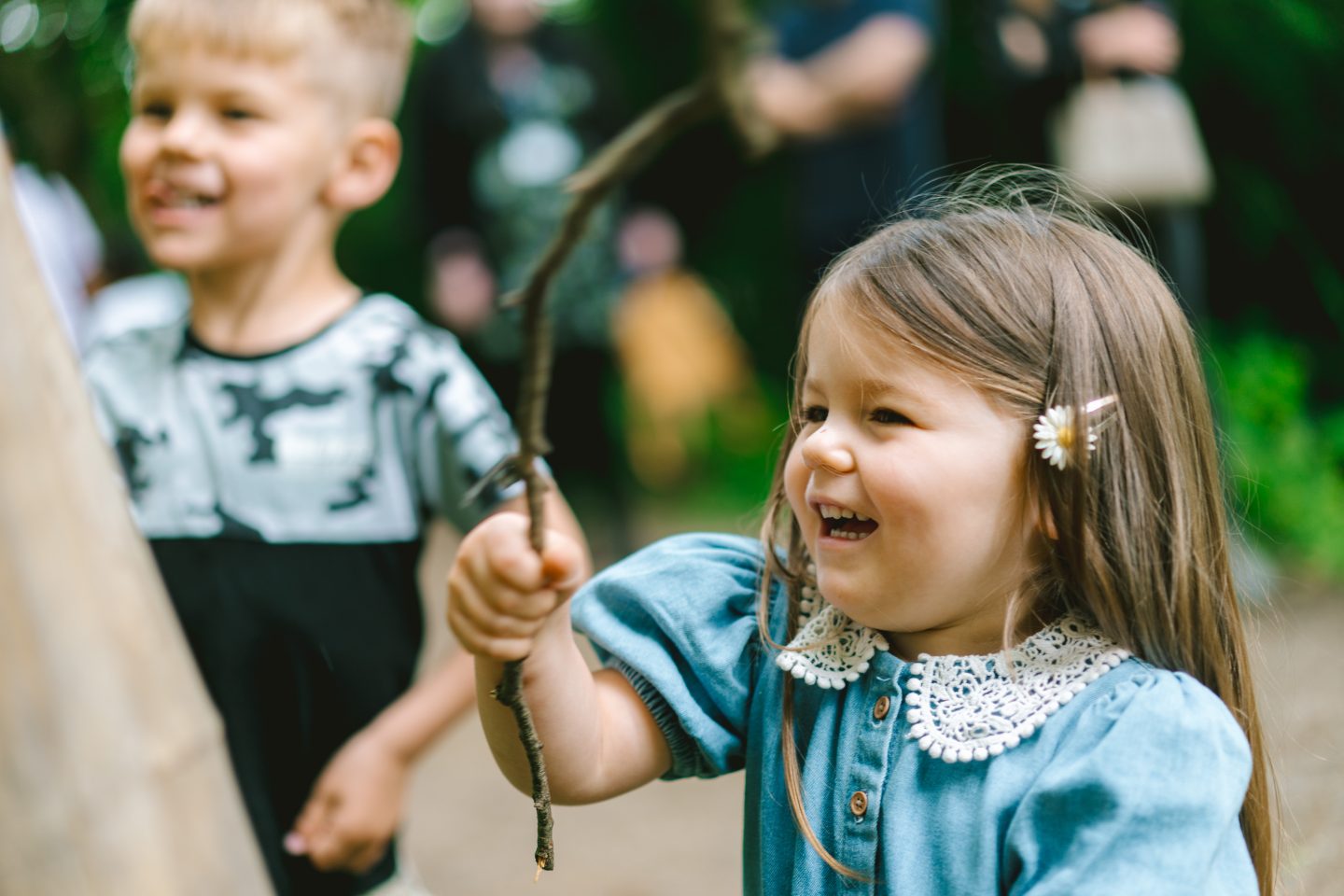 A young girl wearing a denim blue playsuit with a white crochet collar waves a stick she found along the Lakeside path, embracing the spirit of adventure. Surrounded by nature, she explores with curiosity and excitement, discovering the simple joys of the outdoors at Harewood.