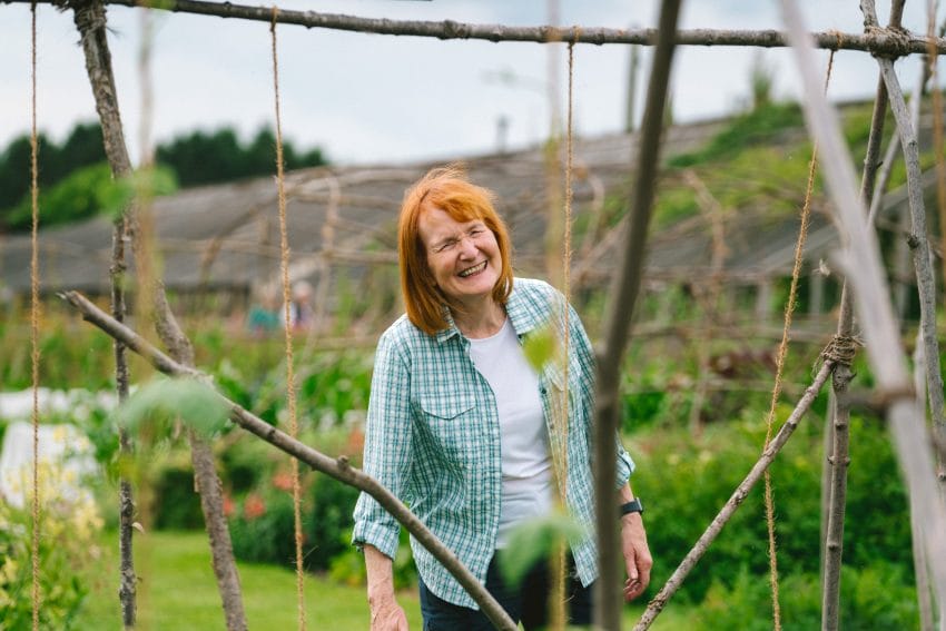 A woman with ginger shoulder-length hair and a fringe smiles as she looks through a structure of garden twine and willow branches, where beans grow below. Surrounded by nature, she enjoys a moment of tranquility, reflecting how Harewood nurtures wellbeing through green spaces, growth, and connection to the outdoors.