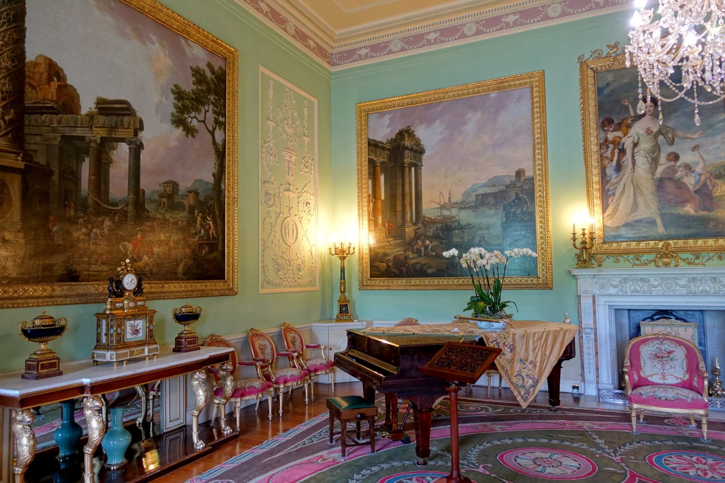 A grand room with high ceilings and a glass chandelier. Gilded Chippendale chairs with pink embroidered fabric surround the room with a dark mahogany piano beside the marble fireplace.
