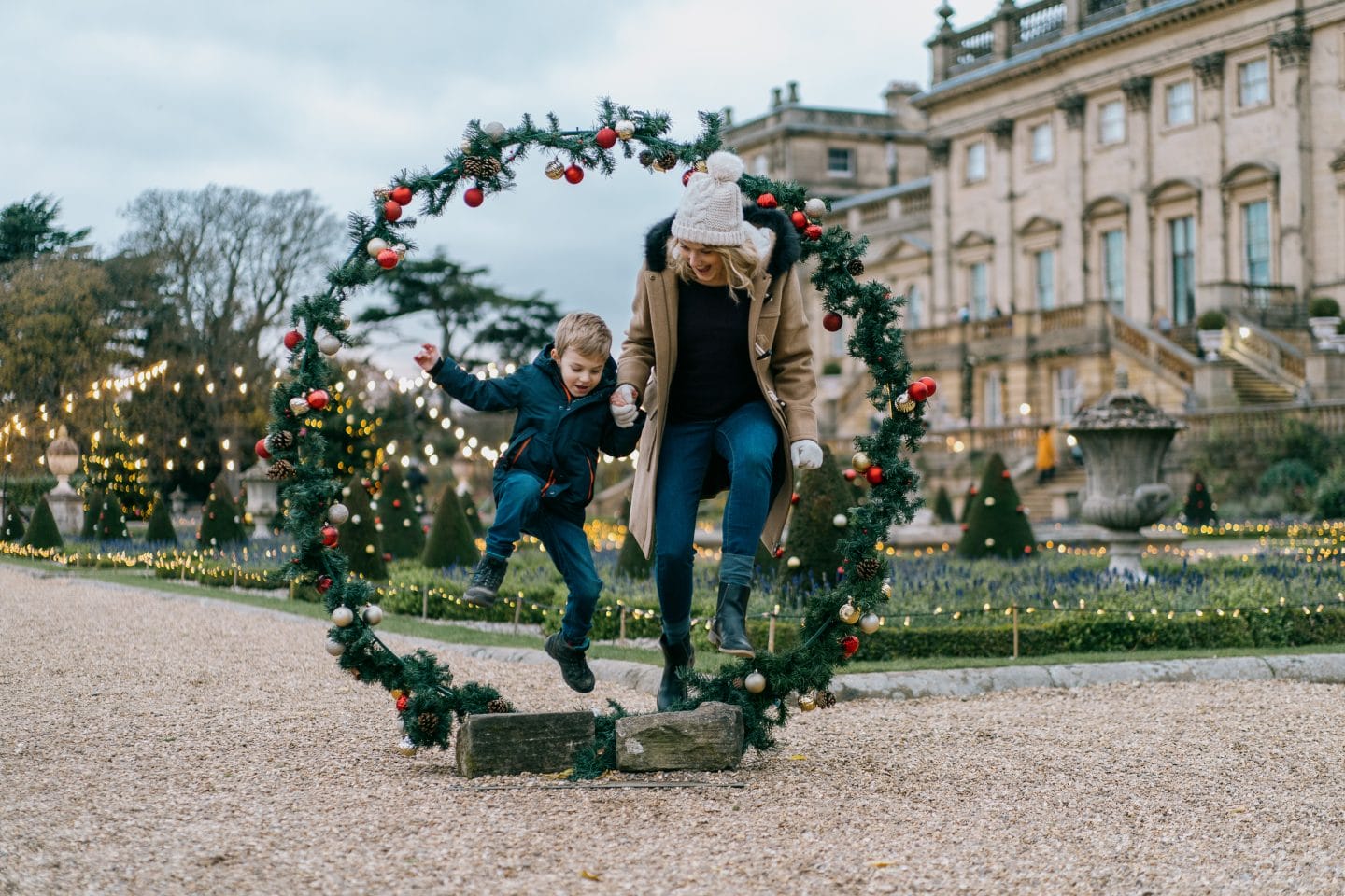 A young women and her son dressed in winter wear gleefully leap through a large wreath decorated with red baubles on the Terrace.