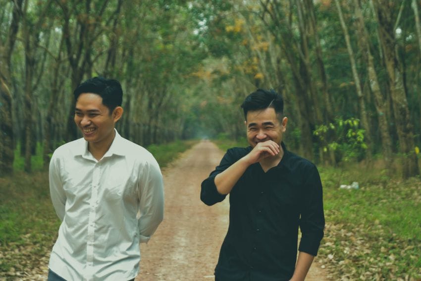 Two men walk down a tree-lined path, smiling and laughing as they enjoy access to Harewood with joint annual Membership. One wears a white shirt, the other black. The scene is relaxed and cheerful.