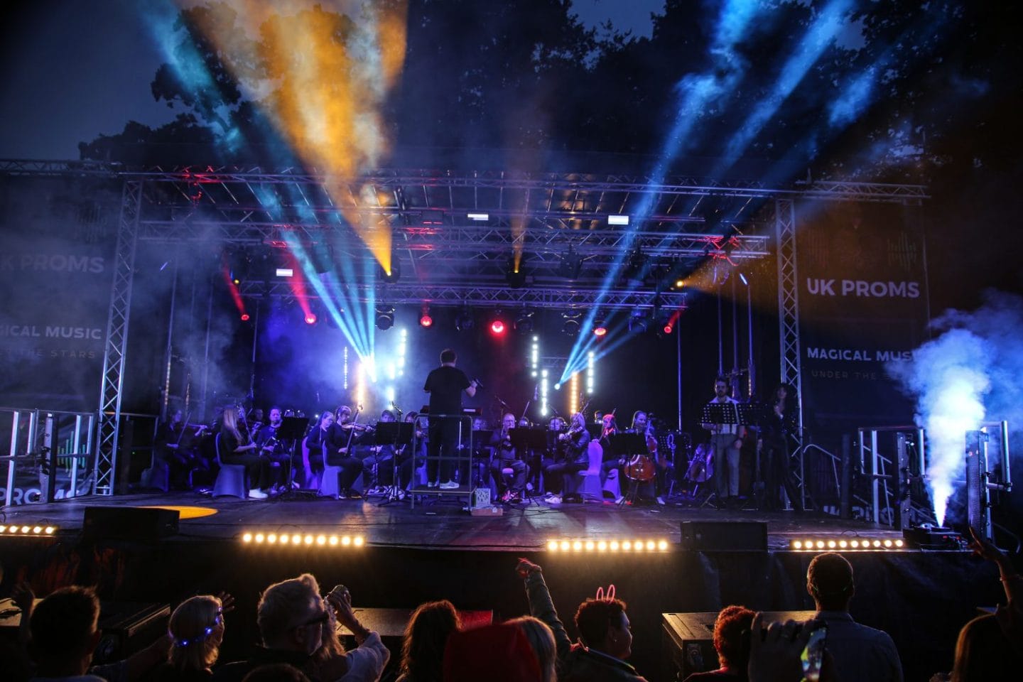 Outdoor concert at night with an orchestra performing on a large, brightly lit stage. Colourful beams of light and mist create a magical, vibrant atmosphere. Audience dances in the foreground.