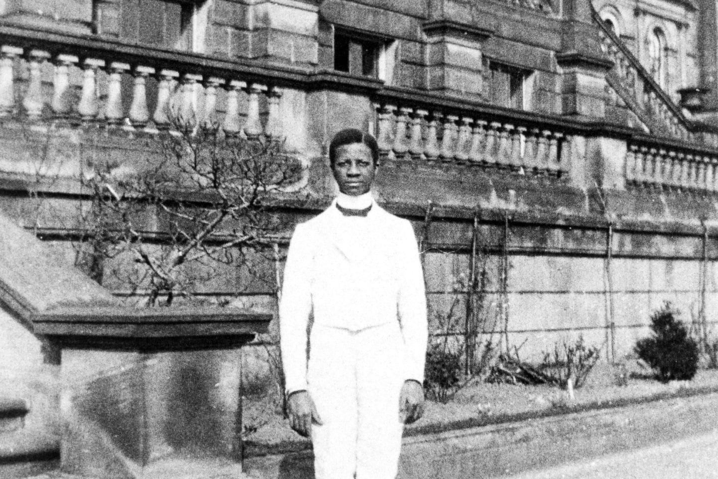 Black and white photo of a man in a white uniform standing outside Harewood House on the Terrace, with stone railings and arched windows, conveying a formal tone.