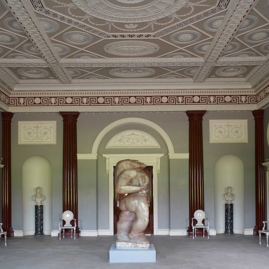A grand room with faux-marble columns lining the walls and an intricate plasterwork ceiling painted in soft pastel colors. In the center, a striking alabaster sculpture of a human figure stands as a focal point. This magnificent space serves as the starting point for exploring Harewood’s historic country house, inviting visitors to step into its rich history, remarkable craftsmanship, and fascinating stories waiting to be discovered.