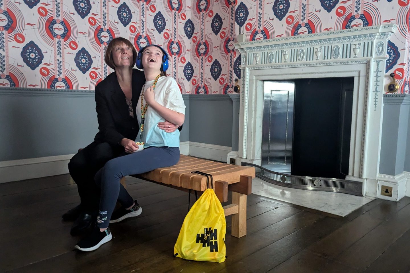 Adult and child sit on a wooden bench in Harewood Watercolour Room looking up at the intricate design of the red and blue hand block printed wallpaper. The child wears a sunflower lanyard and ear defenders. Harewood yellow sensory bag is at the end of the bench.