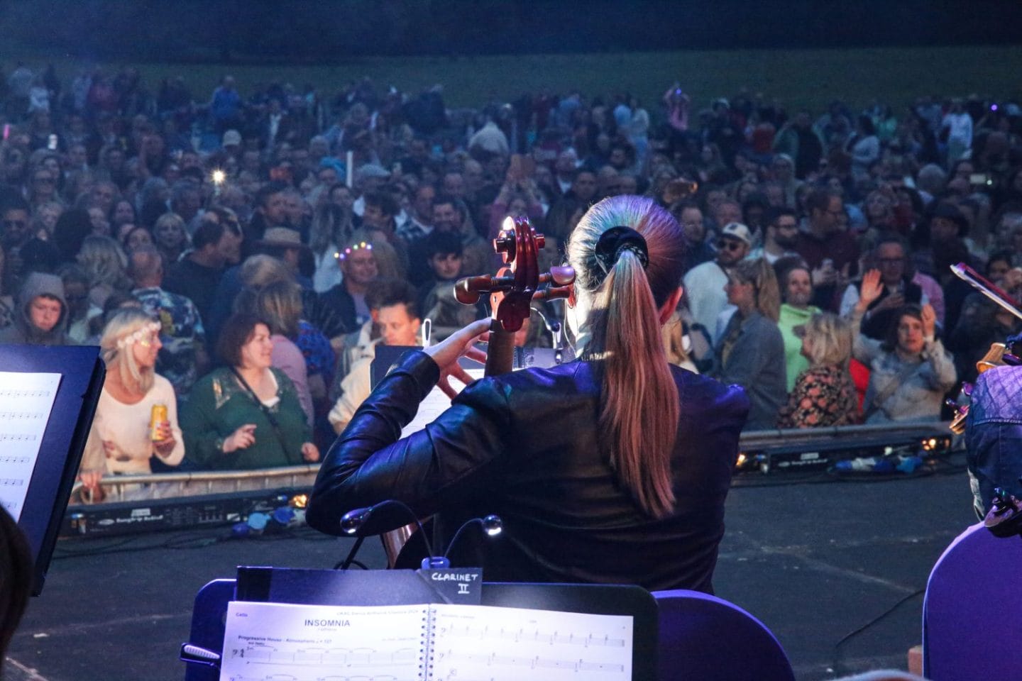 A woman plays the Cello on the stage in front of a large crowd. She has a long ponytail and a black scrunchie in.