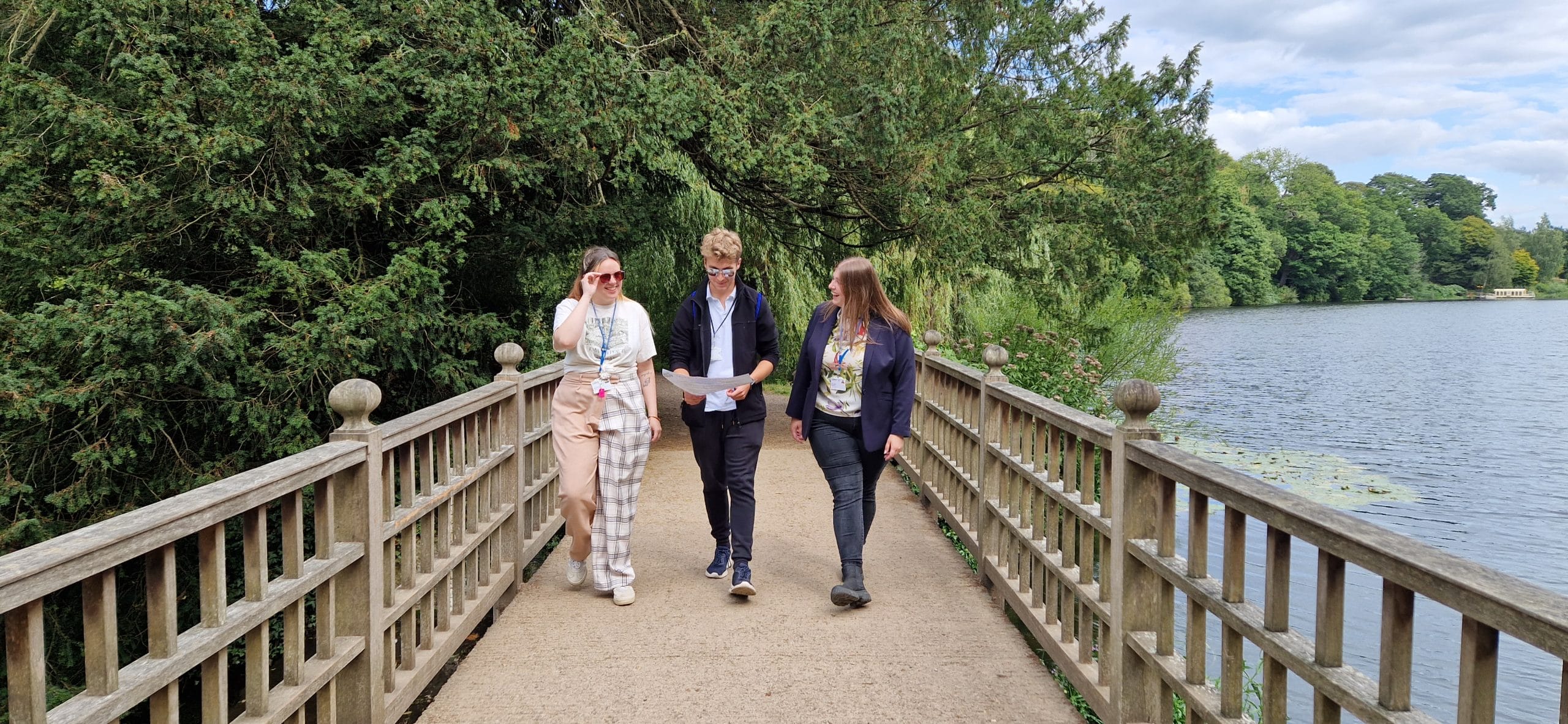 A small group of people hold a scavenger hunt sheet as they walk together over the cascade bridge alongside Harewood lake.