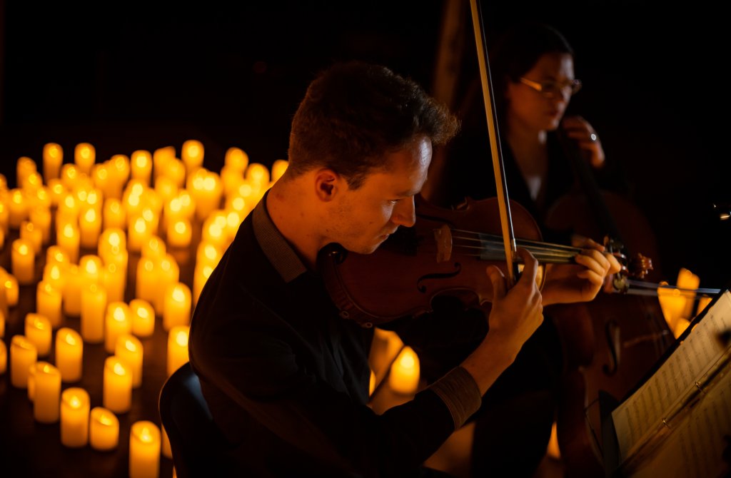 A person plays the violin, illuminated by the warm glow of candles surrounding him. A second person sits behind playing the cello