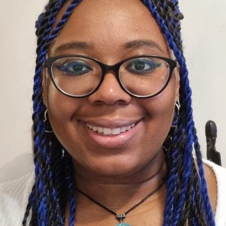 A young women wearing black rimmed glasses with black and blue long braided hair gives a warm and friendly smile to the camera