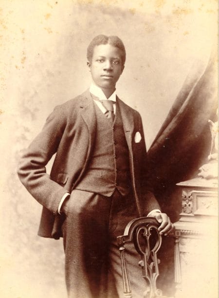A black and white historic photographic portrait of a Black man, George 'Bertie' Robinson, probably in his teens. He is shown three-quarter-length, standing with one arm on a chair and the other in his pocket. He wears a formal tweed-style suit with waistcoat and tie.