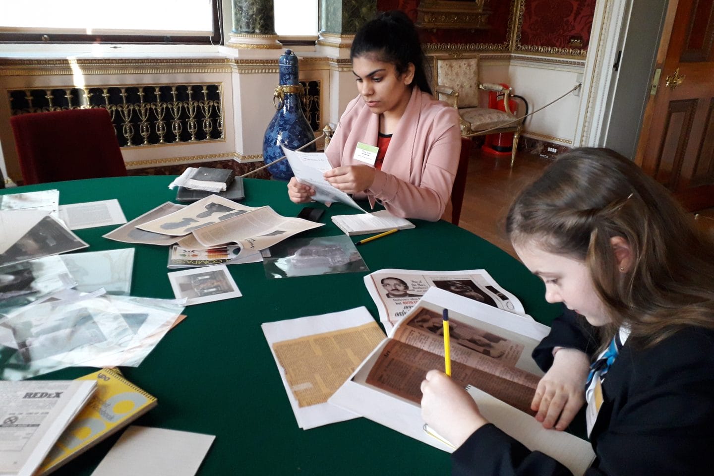 Two young women collaborate at a table in the magnificent Harewood Gallery, focused on their papers, surrounded by books and stationery, deep in study.