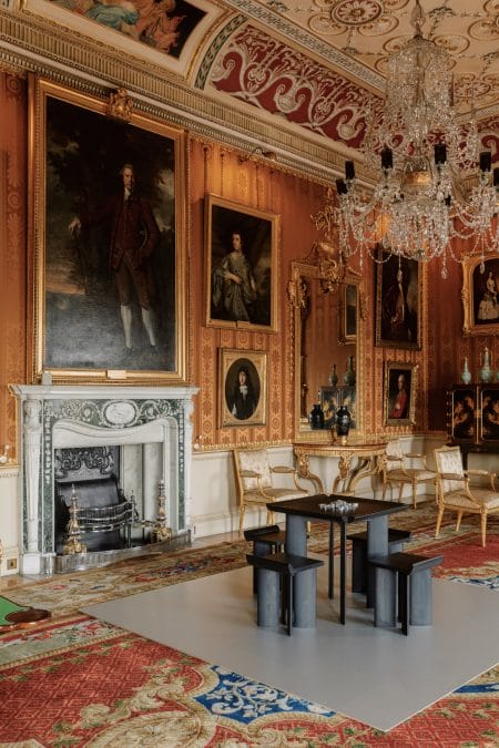 A large historic interior with cinnamon-coloured damask walls and various portraits. In the centre of the room is a black, square-shaped games table with aluminium dominoes.