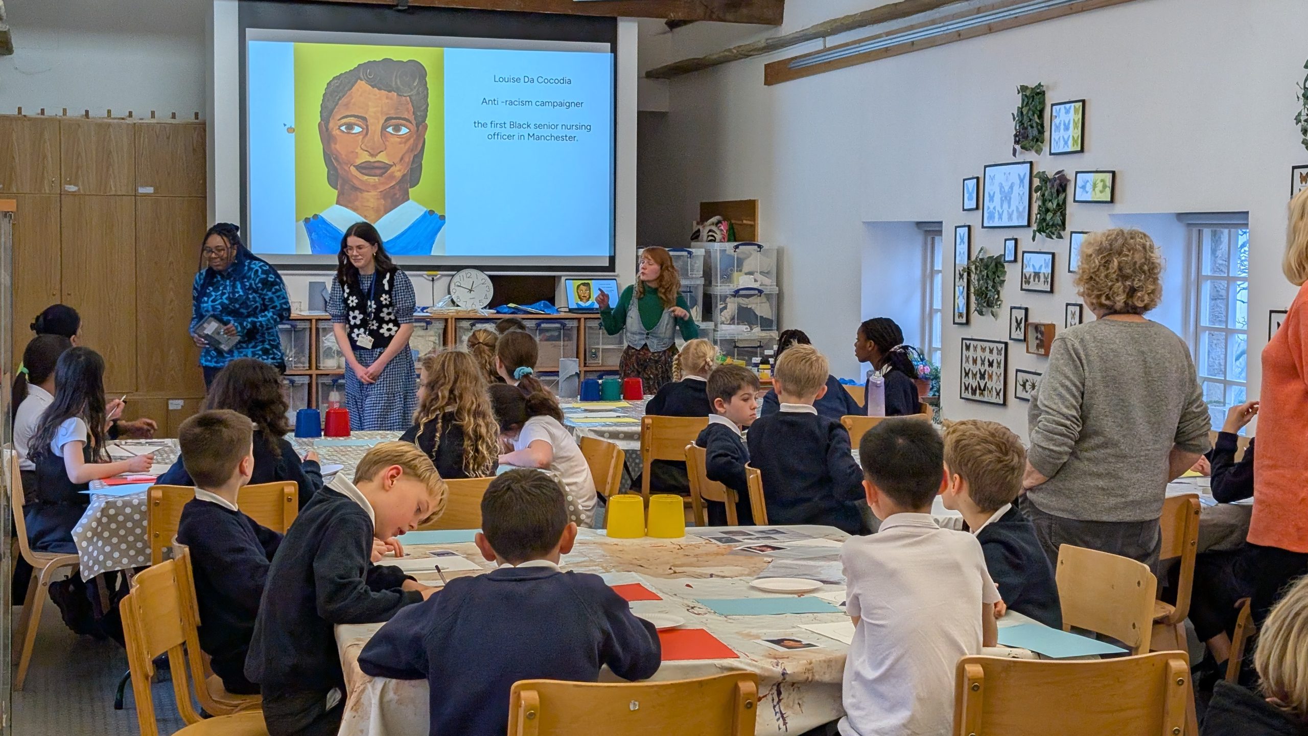 A classroom scene featuring children at tables, attentively listening to a teacher standing at the front.
