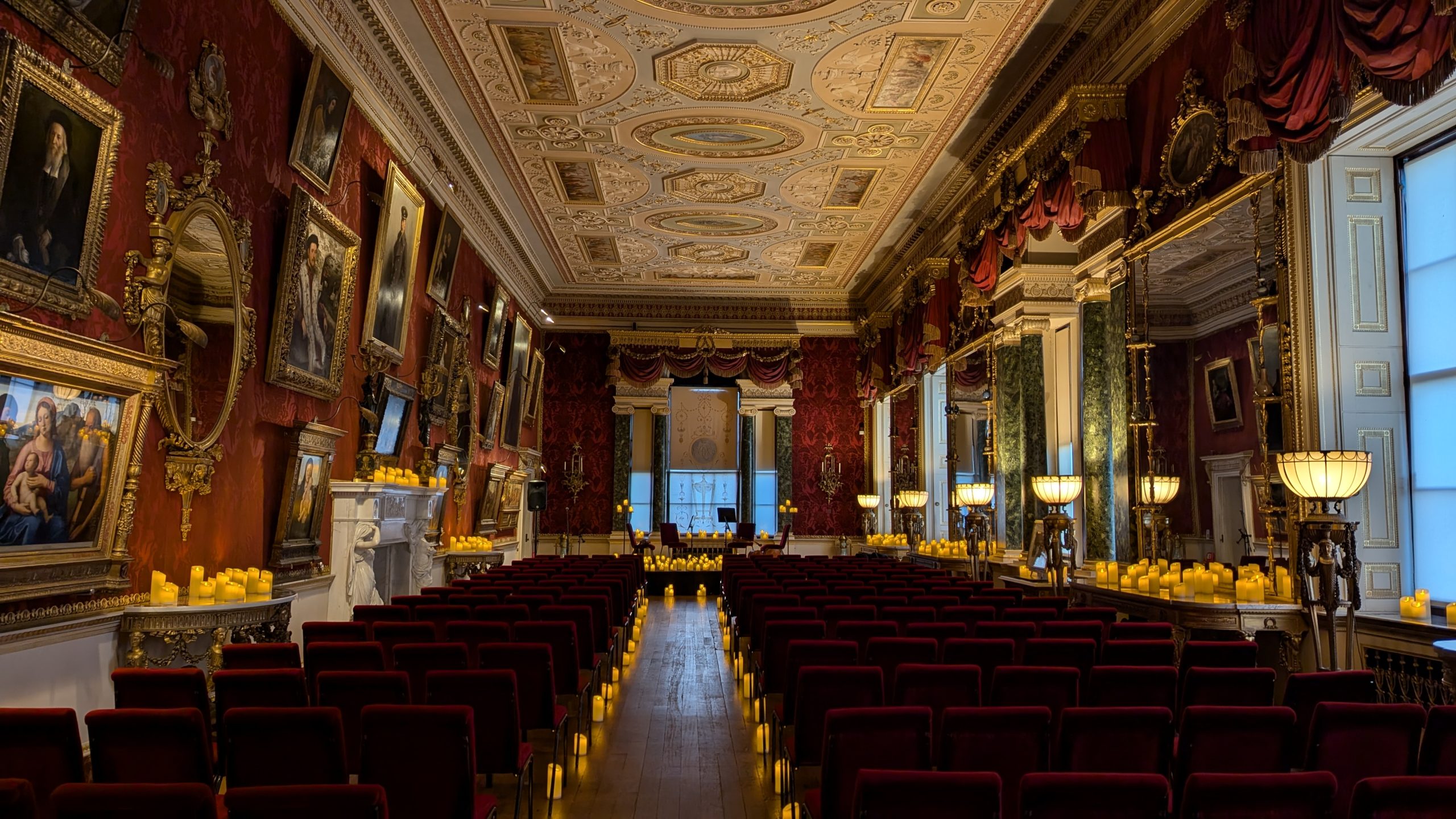 A large, grand room with an ornate ceiling and red silk wallpaper walls. Large mirrors and windows line the right hand side of the room and the left hand wall is full of Renaissance paintings. Rows and rows of red chairs sit facing a stage, with candles lining the isle. The side tables are full of candles