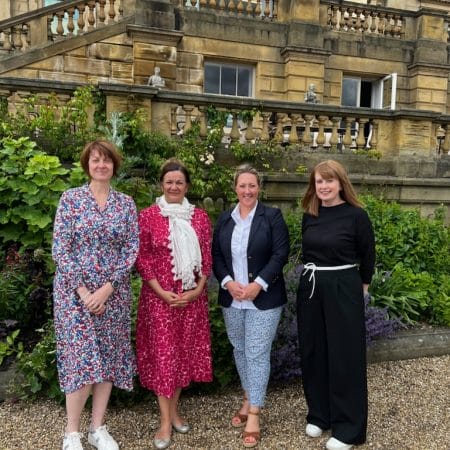 Four women, two in floral dresses, two in suits smile proudly on Harewood Terrace to announce a new partnership between Harewood House Trust and NFU Mutual.