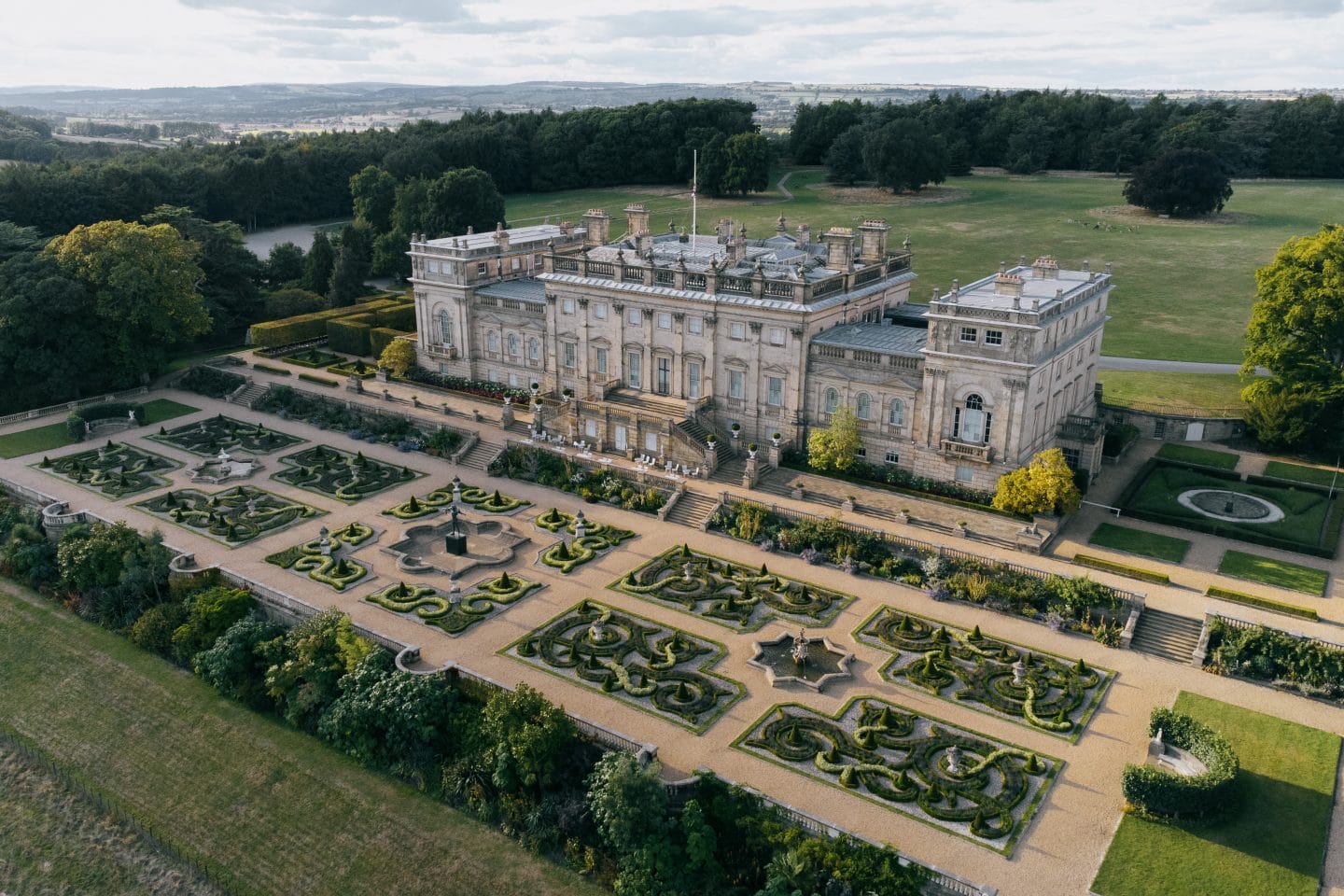 Aerial view of the expansive Harewood House estate, showcasing manicured gardens, intricate hedges, and winding pathways. The historic country house is nestled among vast green fields and trees under a cloudy sky, offering a timeless setting for visitors to explore, unwind, and create lasting memories.