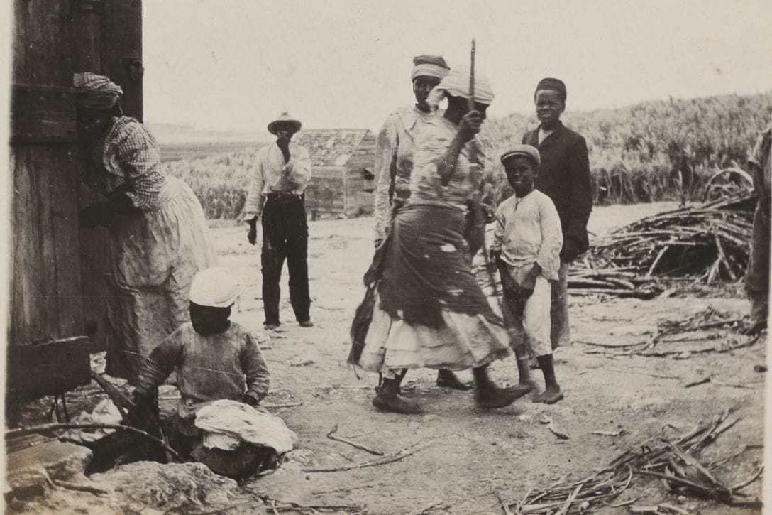 Black and white photograph taken by Countess of Harewood on a trip to Barbados in 1906. A group of people stand outdoors near a wooden structure. Women in headscarves and long skirts are accompanied by children in hats. A man in a hat and another in a dark jacket look on, while the ground is covered with straw and sticks.