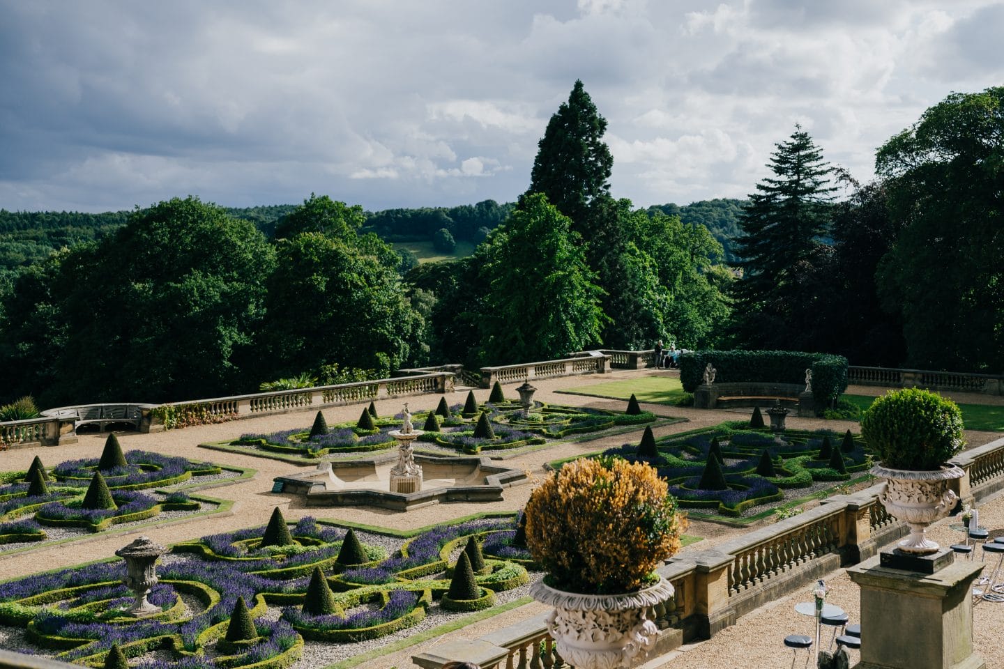 Elegant formal garden with intricate hedges and a central fountain, surrounded by lush green trees under a cloudy sky. Peaceful and serene atmosphere that Harewood Members can enjoy all year round.