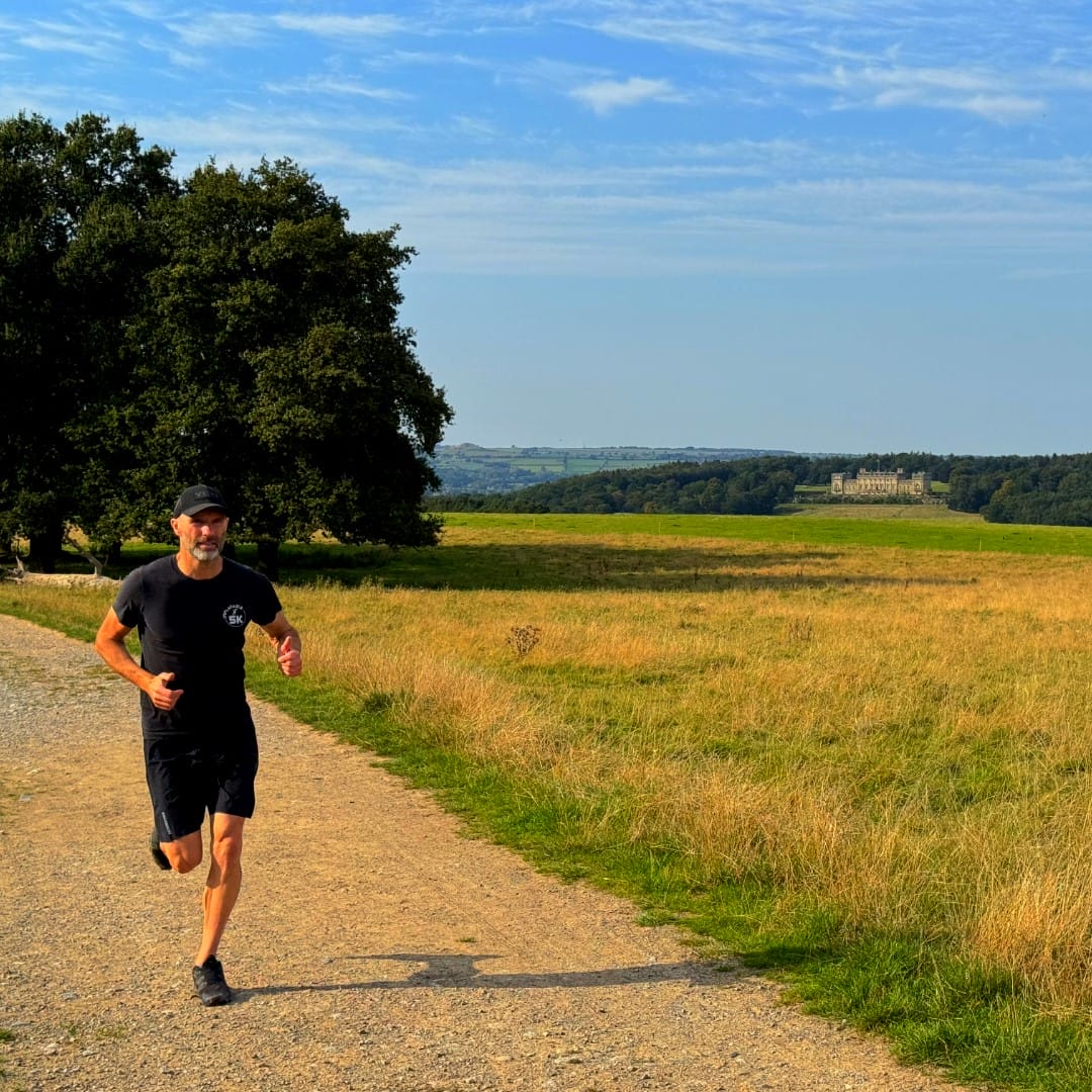 Man running along a country road towards the camera wearing black t-shirt, shorts, and a baseball cap. In the background there is a green field with Harewood House in the distance. The sky is blue.
