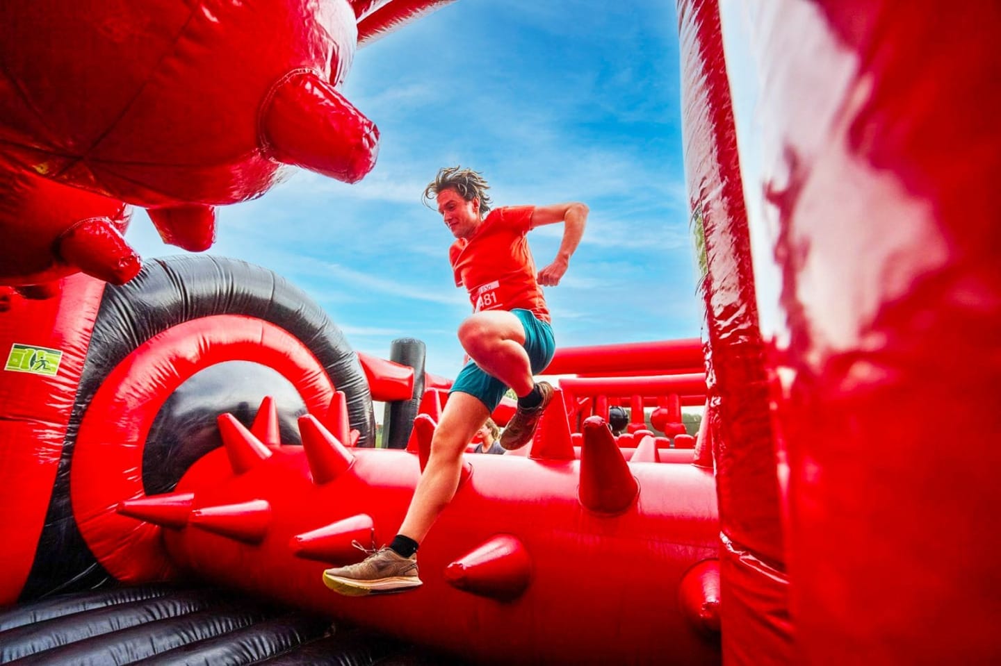Person in athletic clothes jumps over a large red inflatable obstacle course with spikes, under a bright blue sky.