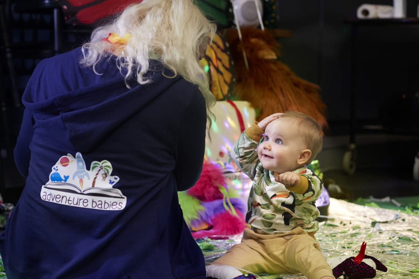 Baby sensory circus session at Harewood House near Leeds. A baby sits on the floor in a colorful play area, smiling and looking up at an adult with long light-colored hair wearing a dark hoodie that says “adventure babies” on the back. The baby has one hand on their head and appears engaged and happy. Around them are bright sensory toys, shiny streamers, and soft colorful objects, creating a playful circus special activity setting.