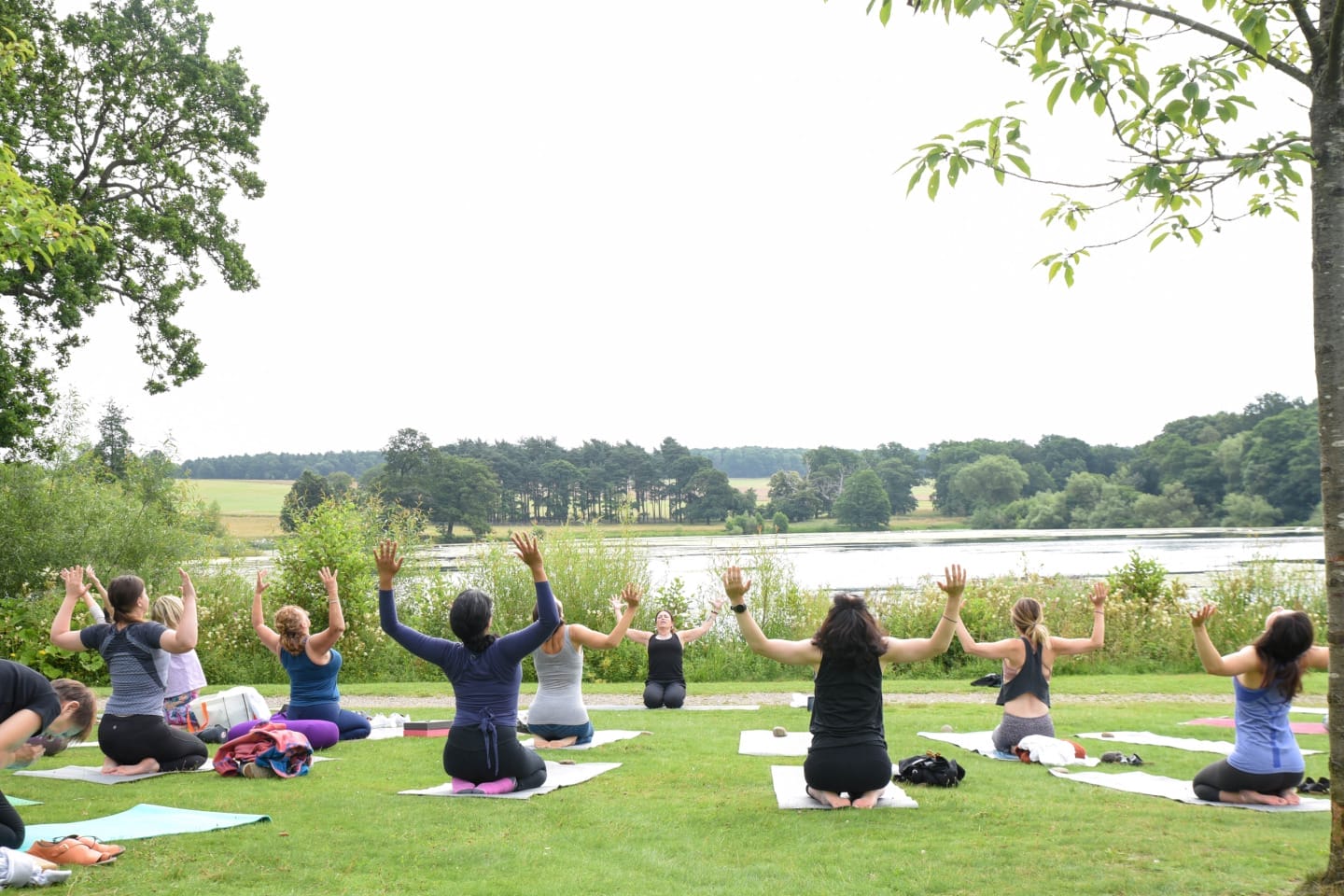 A group of women perform a seated yoga pose with their arms raised towards the sky during an outdoor yoga class on a green lawn beside a Harewood Lake.
