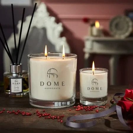 The Dome of Harrogate products stand on a table surrounded by red roses. One large double wick candle, one smaller single wick candle and a reed diffuser. All of which is available to by at Harewood or from our online shop.