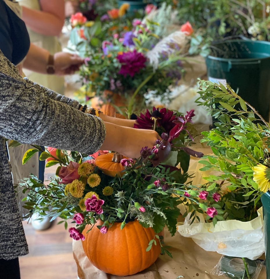 A person wearing a gray sweater is creating a floral centerpiece, placing various flowers into a pumpkin that is being used as a vase. The table is covered with craft paper and scattered flowers, with buckets of flowers in the background.