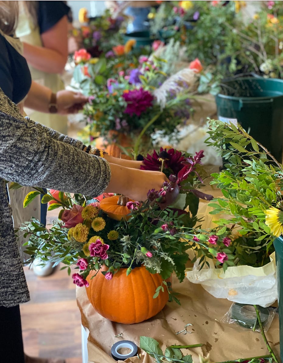 A person wearing a gray sweater is creating a floral centerpiece, placing various flowers into a pumpkin that is being used as a vase. The table is covered with craft paper and scattered flowers, with buckets of flowers in the background.
