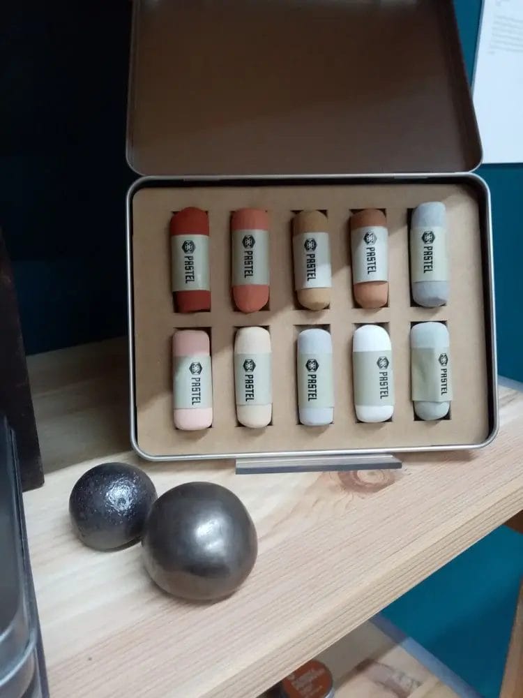 An open metal tin box containing ten hand-rolled soft pastel sticks in various earthy tones and pale shades, arranged neatly in two rows within a cardboard insert.