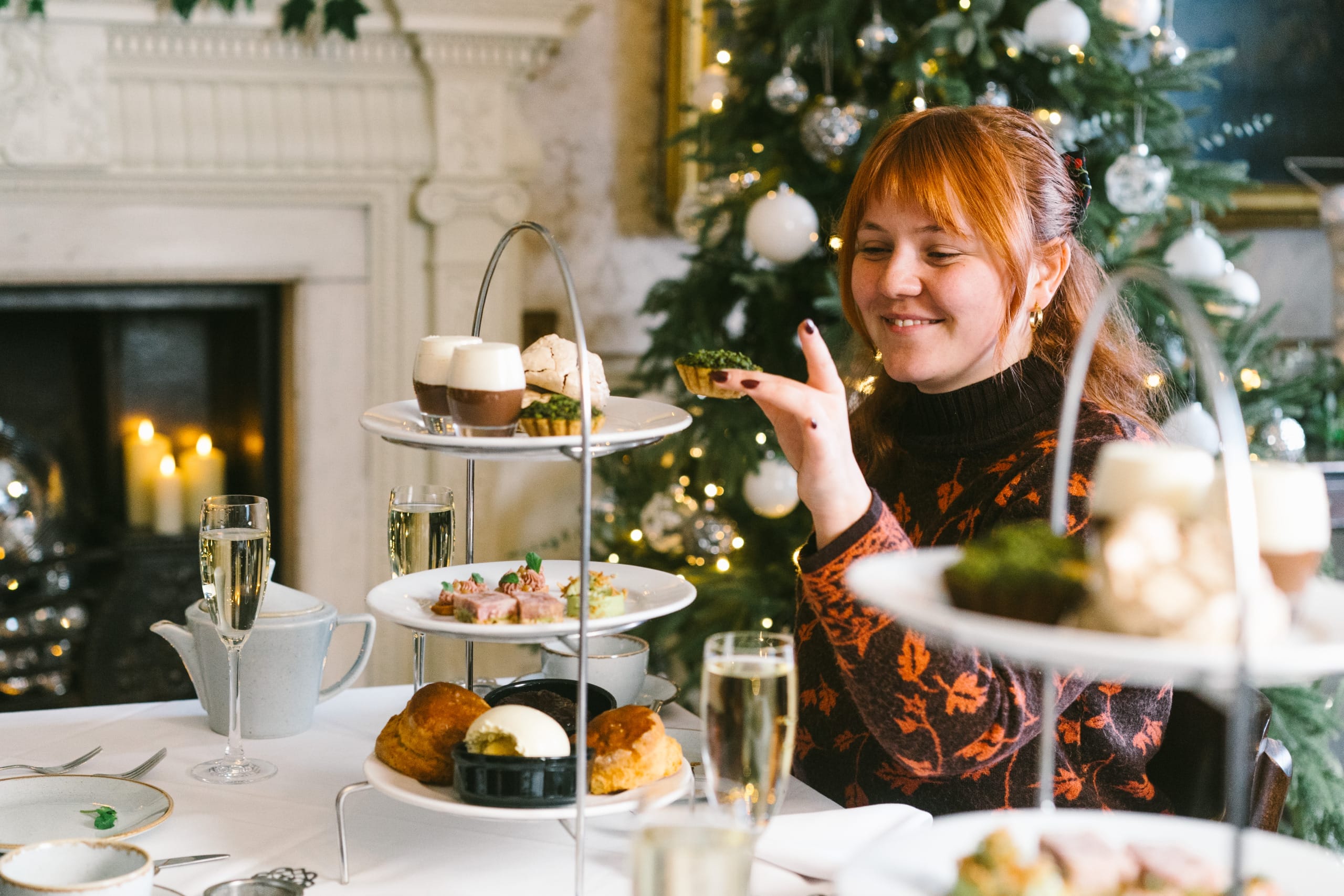 A ginger haired person in her 20's smiles as they select a sweet treat off the top plate of an afternoon tea stand.