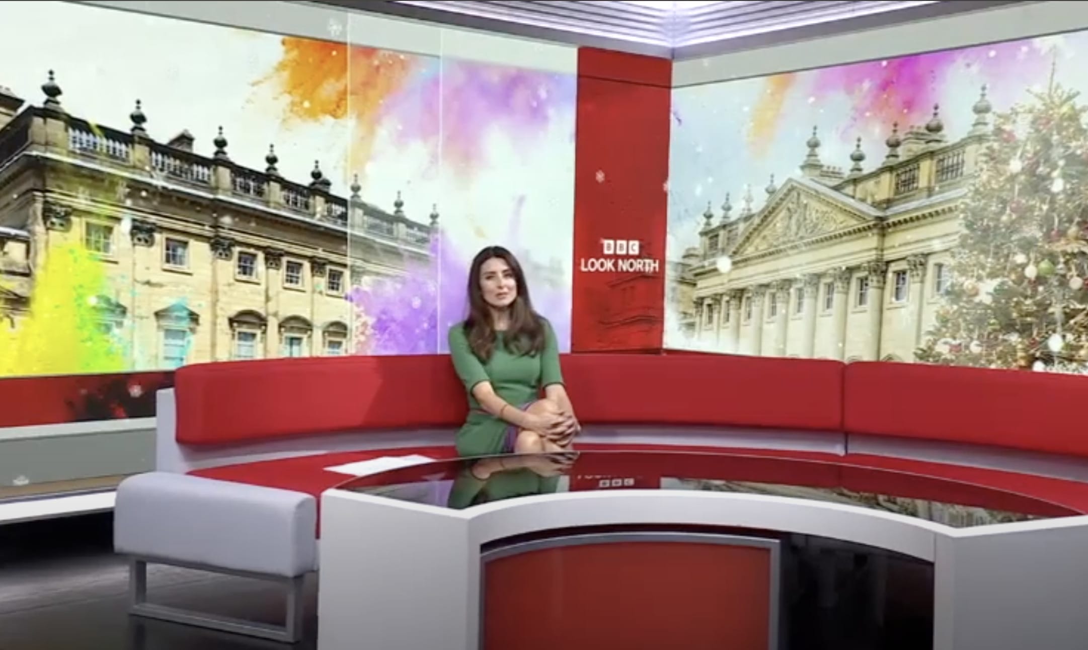 BBC Look North Presenter sits of a red couch in front of a large screen projecting the front of Harewood House.