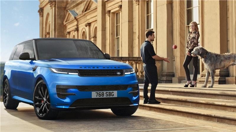Actor Theo James stands next to a large grey wolfhound on the steps of Harewood House Terrace. A bright blue Range Rover Sport is parked on the Terrace just below them.