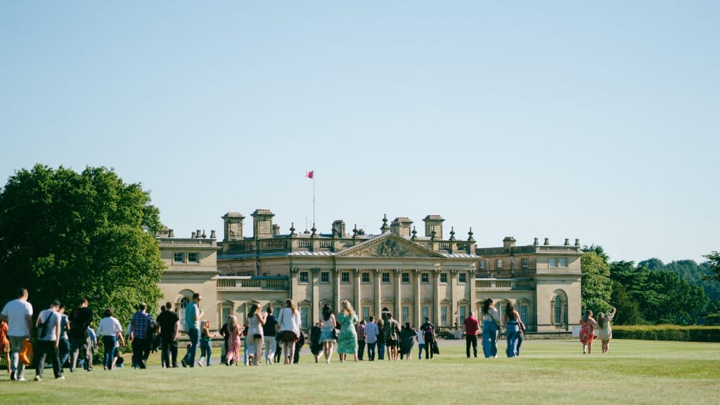 A large group of people walk across the North Front lawn outside Harewood House on a late summers evening.