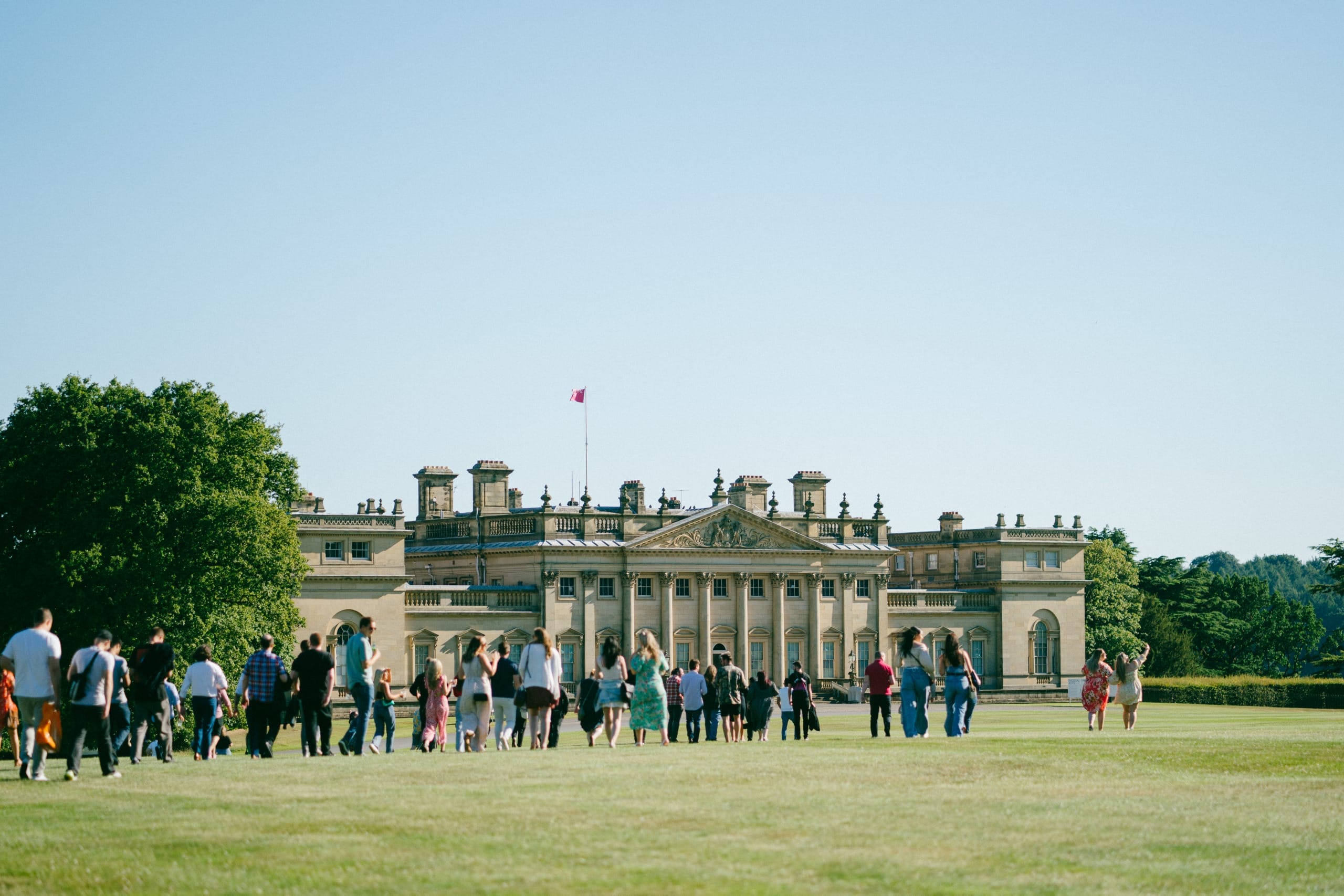 A large group of people walk across the North Front lawn outside Harewood House on a late summers evening.