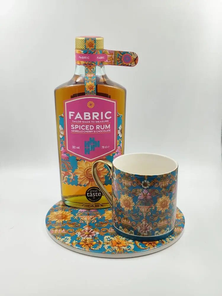 A rectangular bottle of 'Fabric Tailor-Made to Measure Spiced Rum Morello Cherry & Chocolate' with a colourful floral label, placed next to a matching patterned mug and coaster.