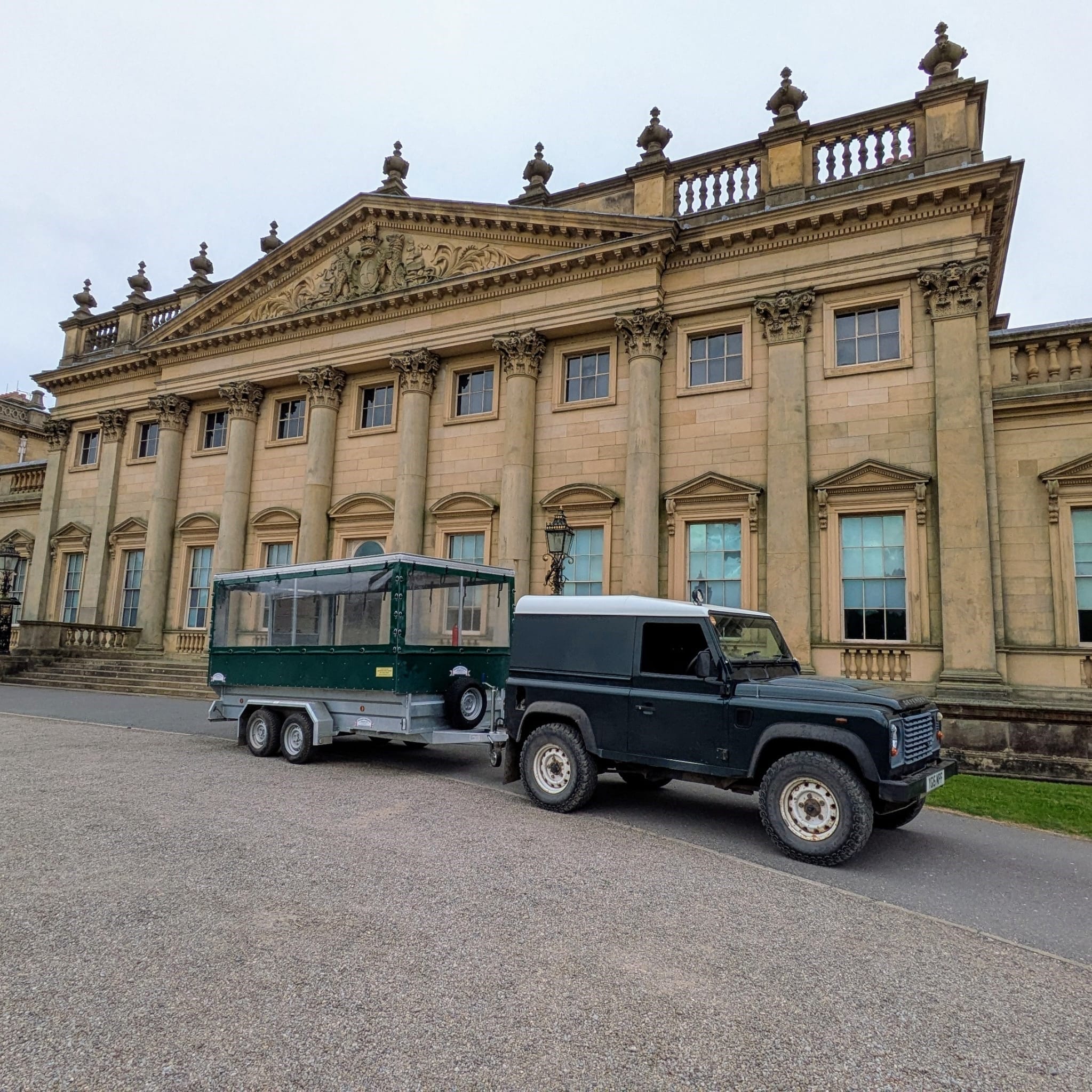 A dark green Land Rover Defender towing a small enclosed trailer is parked on a gravel drive in front of a large, ornate neoclassical stone building with tall columns, decorative carvings, and a balustraded roofline under an overcast sky.