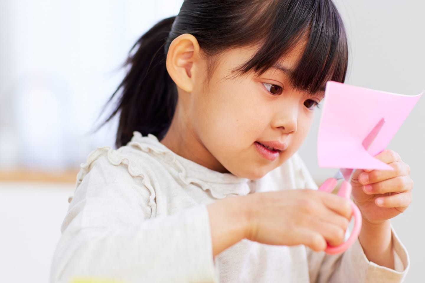 A young girl wearing a white blouse cutting a pink piece of paper with scissors.