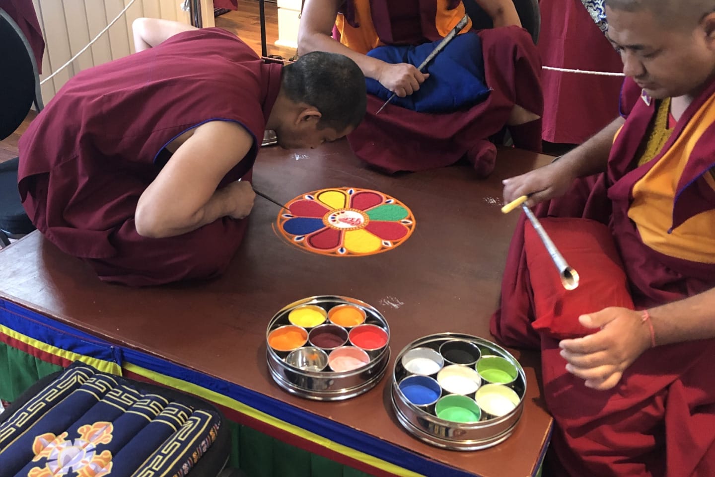 A group of Tibetan Buddhist monks are creating a detailed sand mandala on a low table. One monk is carefully leaning close to the artwork while another uses a metal funnel-like tool (called a chakpur) to place colored sand precisely. Bright pigments in small metal trays are arranged nearby. In the background are ritual objects, a ceremonial drum, and framed portraits, contributing to the monastery or meditation-center setting. The atmosphere appears focused and ceremonial.